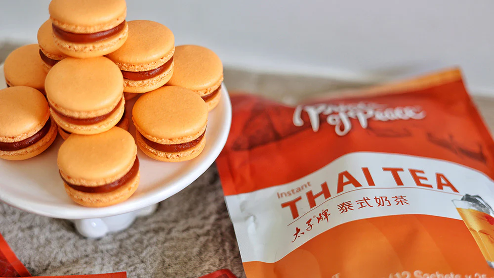 Macaron Thai Tea Elegan: Macaron berwarna jingga cerah dengan isian buttercream Thai Tea, tersusun cantik di atas piring saji